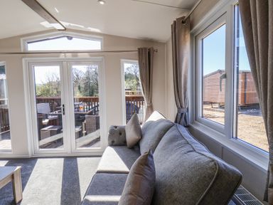 A living area with a grey sofa pillows and glass doors opening to a wooden deck with outdoor chairs and table at Honeysuckle Lodge in Runswick Bay near Staithes