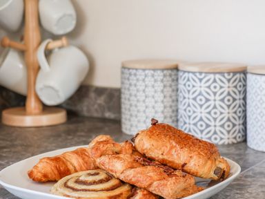 A kitchen countertop with a plate of assorted pastries a wooden mug tree with white mugs and patterned storage jars on the counter at Honeysuckle Lodge in Runswick Bay near Staithes