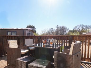 An outdoor deck with a glass top table surrounded by six rattan chairs with cushions and an ice bucket with drinks on the table at Honeysuckle Lodge in Runswick Bay near Staithes