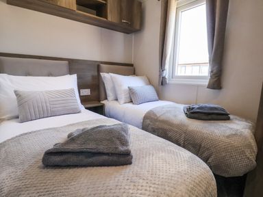 A bedroom with two single beds gray pillows towels and a window with curtains at Honeysuckle Lodge in Runswick Bay near Staithes