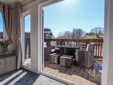 A seating area with a table and chairs on a wooden deck viewed from inside a room with curtains and a cabinet at Lilac Lodge in Runswick Bay near Staithes
