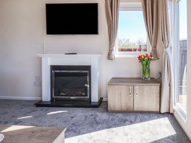 A room with a fireplace a mounted tv a cabinet with flowers by a window and curtains at Lilac Lodge in Runswick Bay near Staithes