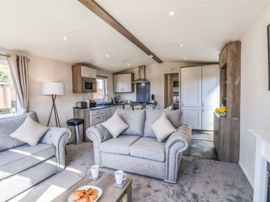 A living room with two grey sofas a coffee table with cups and pastries an open kitchen and a floor lamp at Lilac Lodge in Runswick Bay near Staithes