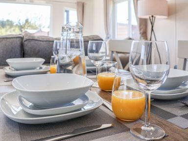 A dining table set with white plates bowls glasses and orange juice in a room with a sofa and windows at Lilac Lodge in Runswick Bay near Staithes