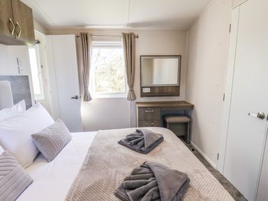 A bedroom with a bed, folded towels on the bed, a window with curtains, a dressing table with a mirror and stool at Lilac Lodge in Runswick Bay near Staithes