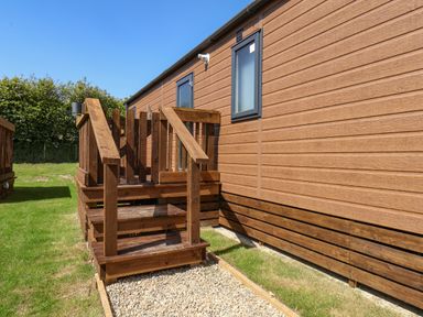 An exterior view of a wooden porch with steps leading to a door on a wooden clad building with grass and bushes surrounding Lilac Lodge in Runswick Bay near Staithes
