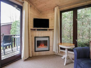 A corner of a room with a wall-mounted TV above an electric fireplace a small round table and a blue armchair near large windows and glass doors at Canadian Cabin 37 in Keswick