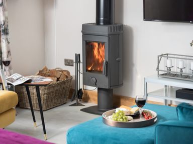 A living room with a wood-burning stove firewood basket and a tray with food and wine on a blue sofa at Shepherds View Dufton near Appleby-in-Westmorland