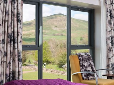 A room with floral curtains a yellow chair with a blanket and a window showing a green hill outside at Shepherds View in Dufton near Appleby-in-Westmorland