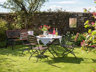 A garden with a stone wall a table with drinks and flowers and wooden chairs at Shepherds View Dufton near Appleby in Westmorland