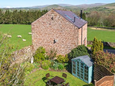 A brick house with a garden shed and patio furniture in a rural setting with sheep in the field at Shepherds View Dufton near Appleby-in-Westmorland