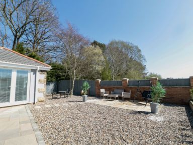 An outdoor patio area with gravel ground two seating areas and trees behind a brick and wooden fence at Old Colliery Cottage in Shilbottle