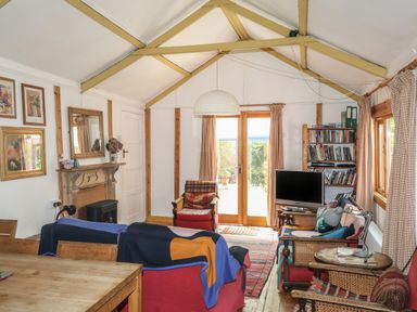 A living room with red armchairs a wooden table a TV on a stand bookshelves and double doors with curtains at Can Yr Afon in Beddgelert