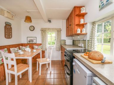 A kitchen with a wooden dining table white chairs wooden cabinets and a loaf of bread on a countertop at Thatch Cottage in Buckland Newton