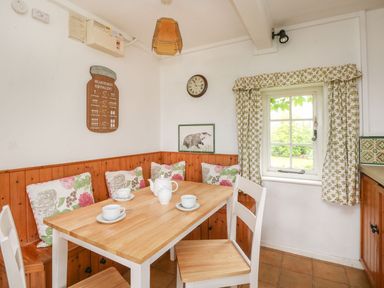 A kitchen nook with a wooden table and white chairs with floral cushions and curtains at Thatch Cottage Buckland Newton
