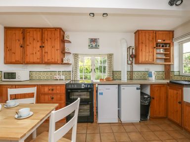 A kitchen with wooden cabinets a black oven a dishwasher a small refrigerator and a wooden table with white chairs at Thatch Cottage in Buckland Newton