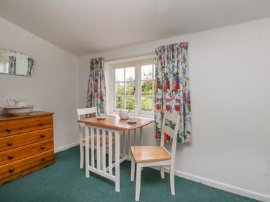 A small dining area with a wooden table and two chairs next to a window with floral curtains and a wooden chest of drawers with a bowl and pitcher on top at Thatch Cottage in Buckland Newton