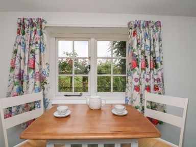 A small wooden table with two white chairs cups and teapot in front of a window with floral curtains at Thatch Cottage in Buckland Newton