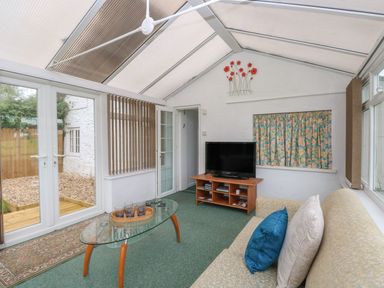 A conservatory with a sofa and pillows a glass coffee table and a television on a wooden stand at Thatch Cottage in Buckland Newton