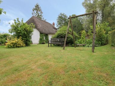 A garden with a wooden swing and bench near a thatched cottage covered in greenery at Thatch Cottage in Buckland Newton