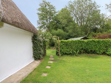 A garden path with stepping stones next to a white thatched building and a wooden archway with greenery at Thatch Cottage in Buckland Newton