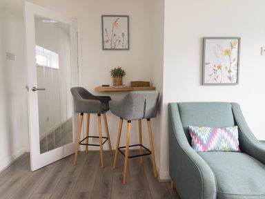 A small dining area with two grey bar stools and a wooden floating shelf table next to a teal armchair with a colorful cushion at The Old Coach House in Conwy