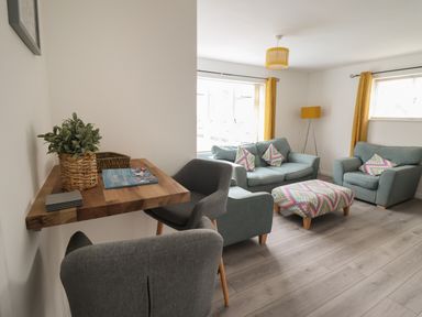 A living room with blue sofas, a patterned ottoman, yellow curtains, a wooden table with chairs, and a plant at The Old Coach House in Conwy