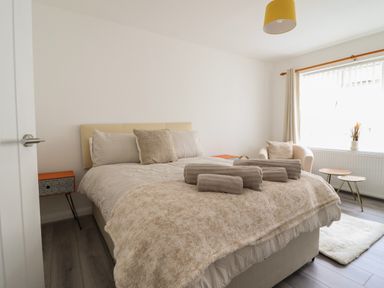 A bedroom with a bed, folded towels on the bed, a bedside table, an armchair, a window with curtains, and a small set of nesting tables at The Old Coach House in Conwy