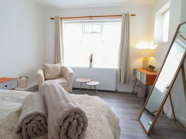 A bedroom with a bed and rolled blankets a beige armchair side tables a wooden dresser with a lamp a standing mirror and a window with curtains at The Old Coach House in Conwy