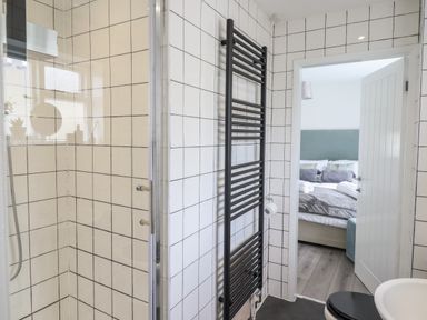 A bathroom with a tiled shower, black towel radiator, toilet, and a view into a bedroom with a bed at The Old Coach House in Conwy