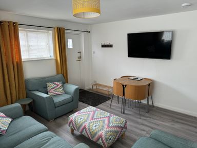 A living room with blue armchairs and sofa a patterned ottoman wooden dining table with chairs yellow curtains and a wall-mounted TV at The Old Coach House in Conwy