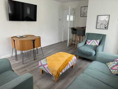 A living room with a sofa armchairs a small table with a yellow throw a wall mounted tv and a small dining table with chairs at The Old Coach House in Conwy