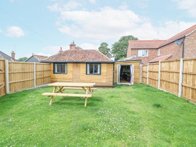 A fenced backyard with a wooden picnic table and a small wooden house with tiled roof and open doors at Squirrel Lodge in North Walsham