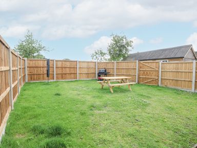A fenced grassy backyard with a wooden picnic table and a barbecue grill at Squirrel Lodge in North Walsham