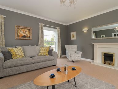 A living room with a gray sofa and patterned cushions a coffee table with cups and a coffee maker an armchair a fireplace and wall art at Cherry Tree House in Highbridge