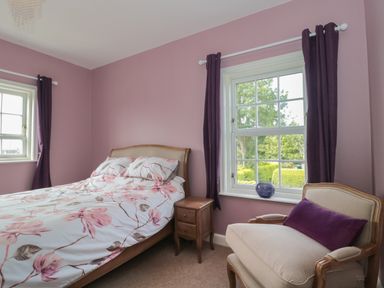 A bedroom with a bed with floral bedding a wooden nightstand two windows with dark curtains an upholstered chair with a purple cushion and a purple vase on the windowsill at Cherry Tree House in Highbridge