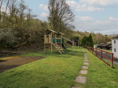 An outdoor playground with wooden climbing structures next to a grassy path and a red wooden fence at Elm Lodge in Keswick