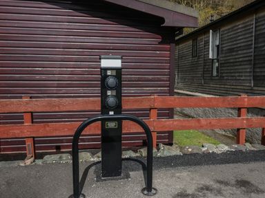 An electric vehicle charging station with two charging sockets in front of a brown wooden building and a red wooden fence