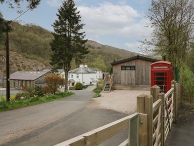 A road with wooden buildings and a red telephone box near trees and hills at Riverside Cottage No 3 in Keswick