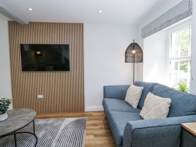 A living room with a blue sofa two cushions a floor lamp a wall-mounted tv and a round table at Riverside Cottage No 3 in Keswick