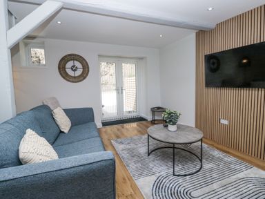 A living room with a blue sofa, round coffee table, wall clock, mounted television, and glass doors at Riverside Cottage No 3 in Keswick