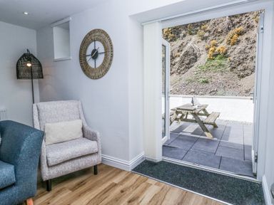 A living room with armchairs clock on wall and open door to patio with picnic table at Riverside Cottage No 3 in Keswick