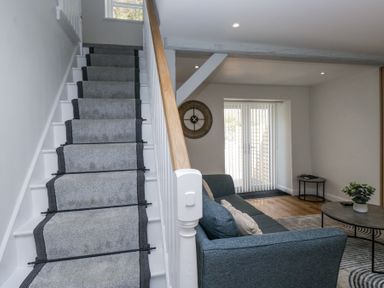 A staircase with carpet runners beside a living area with a sofa, coffee table, side table, clock on the wall and sliding doors at Riverside Cottage No 3 in Keswick