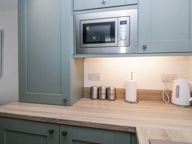 A kitchen countertop with a microwave oven above and canisters labeled tea coffee sugar beside a paper towel roll and electric kettle at Riverside Cottage No 3 Keswick