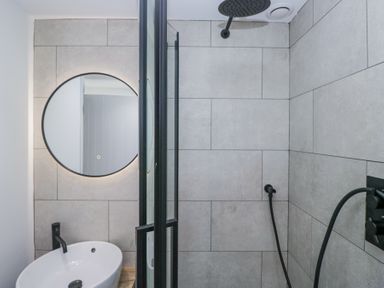 A bathroom with a round mirror above a white sink and a walk-in shower with gray tiles