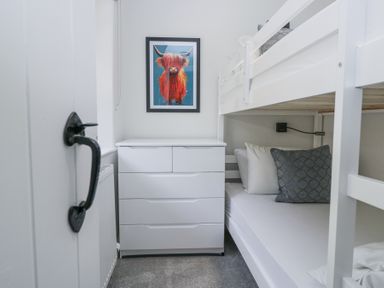A small bedroom with white bunk beds a white chest of drawers and a framed picture of a red highland cow on the wall at Riverside Cottage No 3 in Keswick