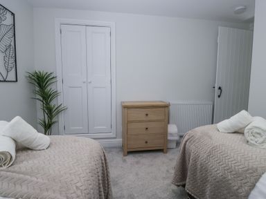 A bedroom with two beds covered with beige blankets and white towels on top a wooden chest of drawers a white closet door a plant and an open door at Riverside Cottage No 3 Keswick