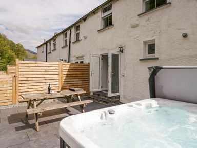 An outdoor patio with a wooden picnic table and open double doors beside a bubbling hot tub at Riverside Cottage No 3 in Keswick