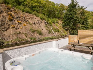 An outdoor hot tub with bubbling water and a wooden picnic table with a bottle and glasses near a hillside at Riverside Cottage No 3 in Keswick