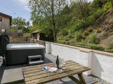 An outdoor patio with a hot tub a wooden picnic table with wine and glasses next to a white wall and trees at Riverside Cottage No 3 in Keswick